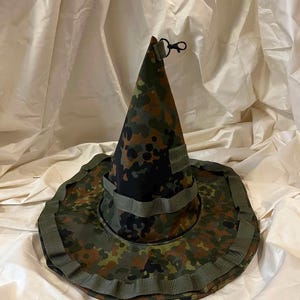 Può includere: Un cappello conico con motivo mimetico nei toni del verde, marrone e nero. Il cappello presenta una tesa larga con passanti in fettuccia verde oliva integrati e una piccola clip in plastica nera fissata sulla punta della calotta.