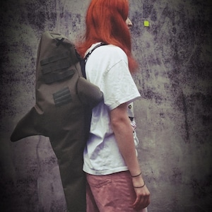 Könnte beinhalten: Ein großer, olivgrüner Rucksack in Haiform mit Rückenflosse, Seitenflossen und Schwanzflosse. Der Rucksack verfügt über schwarze Träger und horizontale schwarze Gurtbanddetails an der Seite. Er wird über einem weißen T-Shirt und einer rosa Hose getragen.