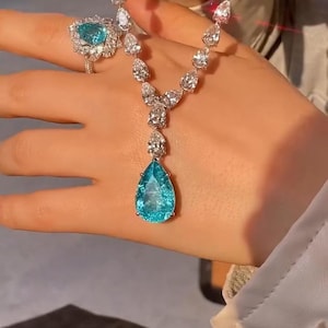 May include: A silver-toned necklace and ring set featuring pear-shaped, bright turquoise-blue gemstones surrounded by smaller, clear, sparkling white stones. The necklace has a central pear-shaped pendant, and the ring features a matching stone.