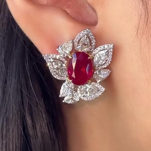 Puede incluir: Un pendiente con forma de flor que presenta un gran rubí rojo intenso de talla ovalada en el centro, rodeado por múltiples diamantes blancos de talla pera y redonda engastados en un marco metálico, creando un diseño de racimo floral.