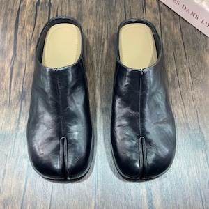 May include: A pair of black slip-on mules featuring a distinct split-toe design at the front. The shoes have a smooth, dark exterior finish and a light beige interior footbed. The construction includes a central seam running from the toe split.