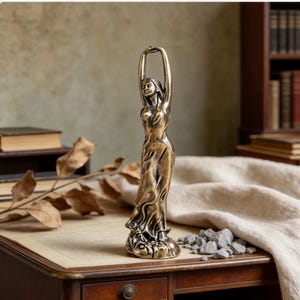 Peut inclure: Une figurine en bronze représentant une personne debout, les bras levés au-dessus de la tête. Le personnage porte un long vêtement fluide avec des plis et des drapés visibles, posé sur une base texturée. La finition métallique a des reflets sombres.