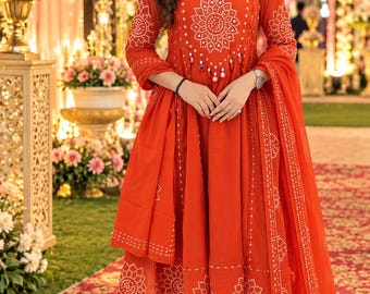 May include: A vibrant orange long-sleeved kurta dress featuring a white circular mandala-style print, a keyhole neckline, and a matching sheer dupatta with white dotted patterns. The outfit includes slim-fit trousers and is paired with open-toe sandals.