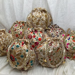 以下が含まれることがあります： ゴールド、レッド、ブルー、グリーンの複雑な花柄刺繍が施されたクリーム色の生地の巾着袋のコレクション。各ポーチには、白いフェイクパールとゴールドトーンの金属製アクセントで作られた装飾的なハンドルが付いています。