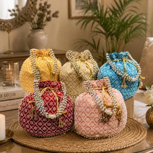 Puede incluir: Cinco bolsas redondas con cordón en amarillo, dorado, azul, rojo y melocotón, cada una con intrincados bordados florales blancos y un asa decorativa de perlas artificiales blancas. Las bolsas tienen cierres de cordón dorados con borlas.