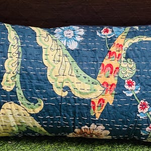 Puede incluir: Un cojín rectangular con fondo azul oscuro y un estampado floral y de hojas multicolor en tonos amarillo, verde claro, rojo y blanco. La tela muestra detalles visibles de acolchado con puntadas horizontales de color blanco.