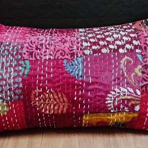 May include: A rectangular throw pillow featuring a patchwork design in shades of magenta, deep pink, and red. The fabric is adorned with various floral and paisley patterns in white, gold, and blue, accented by visible white running stitch embroidery.