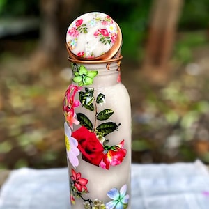 Könnte beinhalten: Eine zylindrische Metallwasserflasche mit hellbeiger Oberfläche, verziert mit einem lebendigen Blumenmuster aus roten Rosen, grünen Blättern und verschiedenen kleinen bunten Blüten. Die Flasche hat einen passenden runden Deckel.