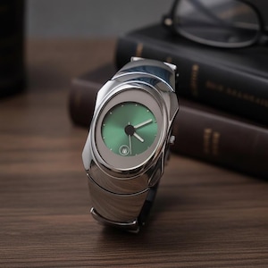 Könnte beinhalten: Eine Armbanduhr mit einem polierten silberfarbenen Metallgehäuse und einem runden grünen Zifferblatt. Die Uhr verfügt über silberfarbene Stunden- und Minutenzeiger, einen zentralen schwarzen Sekundenzeiger und ein kleines Datumsfenster bei sechs Uhr.