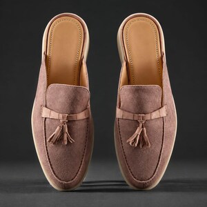 May include: A pair of mauve suede mule loafers featuring a pointed toe, a decorative strap with a matching tassel detail across the vamp, and a light tan interior lining. The shoes have a flat sole and a slip-on design with an open back construction.