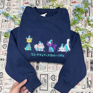 以下が含まれることがあります： とんがり帽子をかぶった4匹の猫の刺繍が施されたネイビーブルーのクルーネックトレーナー。猫の下には、明るいティール色の糸で日本語のテキスト「ココ・テティア・アガット・リチェ」が刺繍されています。トレーナーは長袖で、袖口と襟元はリブ編みになっています。