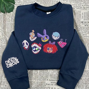 Könnte beinhalten: Ein marineblaues Sweatshirt mit Rundhalsausschnitt, das auf der Brust einen Grafikdruck verschiedener bunter Zeichentrickfiguren und auf dem rechten Ärmel den weißen Schriftzug "THE AMAZING DIGITAL CIRCUS" aufweist. Langarm mit Rippbündchen.
