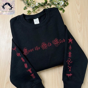 May include: A black long-sleeve crewneck sweatshirt featuring red text on the chest that reads Fear the Old Blood. The sleeves are decorated with a series of red vertical symbols and geometric designs printed along the length of each arm.