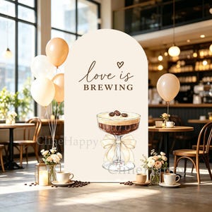May include: A tall, arched, cream-colored display sign featuring the brown text "love is BREWING" with a small heart icon. Below the text is a detailed illustration of a cocktail glass filled with dark liquid, topped with coffee beans and a bow.