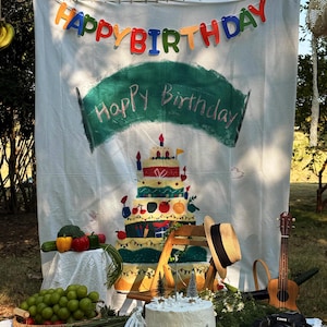 Könnte beinhalten: Ein weißer Stoffhintergrund mit einem bunten "HAPPY BIRTHDAY"-Banner, einem bemalten grünen Banner mit dem Text "Happy Birthday" und einer Illustration einer mehrstöckigen Torte. Darunter stehen eine weiße Torte, Weintrauben, Erdbeeren, eine Ukulele und eine Kamera.