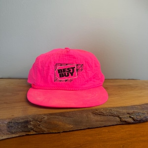 May include: A bright pink baseball cap with a flat brim. The cap features the text "BEST BUY" in black, within a black and white geometric design. The hat is made of a nylon-like material and sits on a wooden surface.