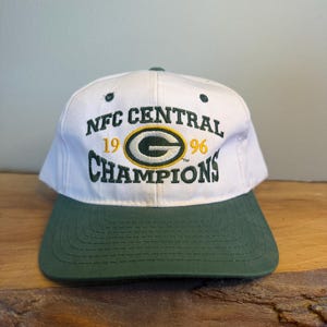 Puede incluir: Gorra de béisbol blanca y verde con el texto "NFC CENTRAL CHAMPIONS 1996". La gorra presenta el logotipo de los Green Bay Packers en el centro. La visera es verde y la corona es blanca.
