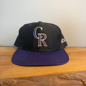 May include: Black baseball cap with a purple peak. The cap displays the Colorado Rockies logo in purple and white embroidery. The hat is on a wooden surface.