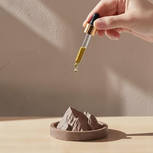 Puede incluir: Un cuentagotas de cristal con cuello dorado y bulbo negro que dispensa una gota de líquido dorado sobre una pequeña escultura circular de color marrón con forma de montaña. La escultura presenta picos dentados detallados y una base plana.