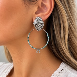 Peut inclure: Une boucle d'oreille argentée composée d'un clou en forme de coquillage texturé relié à un grand anneau. L'anneau est orné de petites perles turquoise et argentées et comporte une breloque en fer à cheval avec des pierres claires.