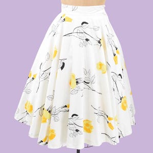 Puede incluir: Falda blanca acampanada con un vibrante estampado floral. El diseño presenta rosas amarillas y tallos y hojas dibujados en negro. La falda se exhibe en un maniquí sobre un fondo lavanda.