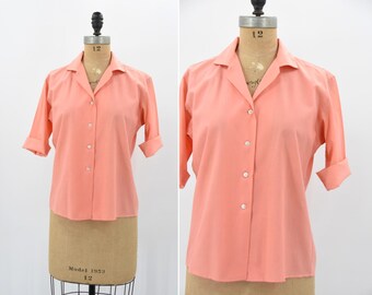 Puede incluir: Una camisa de color melocotón con botones, cuello y mangas tres cuartos. La camisa está hecha de una tela ligera y tiene un corte holgado.