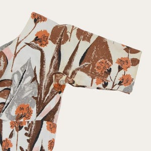 May include: Close-up of a patterned fabric featuring a floral design. The fabric has a white background with brown, orange, gray, and pink floral and leaf patterns. The sleeve of a garment is visible, with a button closure.