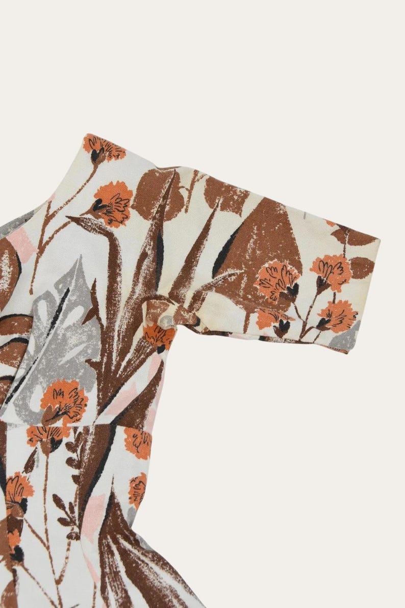 May include: Close-up of a patterned fabric featuring a floral design. The fabric has a white background with brown, orange, gray, and pink floral and leaf patterns. The sleeve of a garment is visible, with a button closure.