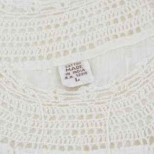 May include: Close-up of a white cotton top with intricate crochet detailing around the neckline. The tag reads "Cotton Made in India" and indicates a size "L". The top has a delicate, airy appearance.
