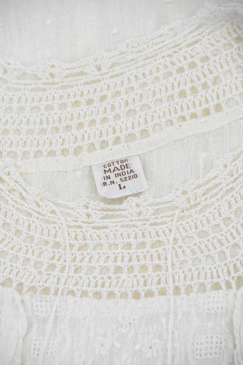May include: Close-up of a white cotton top with intricate crochet detailing around the neckline. The tag reads "Cotton Made in India" and indicates a size "L". The top has a delicate, airy appearance.