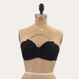 May include: A black strapless top featuring a large, structured bow design across the front. The garment is made of a solid black fabric with gathered pleats that create a voluminous, layered appearance. It is displayed on a beige dress form.