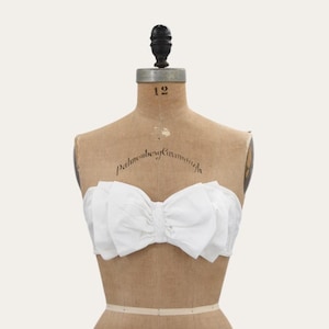 May include: A strapless white top featuring a large, layered bow design across the front. The garment is displayed on a vintage-style beige fabric dress form with the number 12 printed on the neck and cursive text across the chest area.