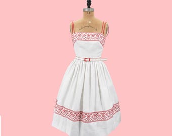 May include: A white dress with red embroidered hearts around the neckline and hem. The dress has a fitted bodice and a full skirt. It is cinched at the waist with a white belt.