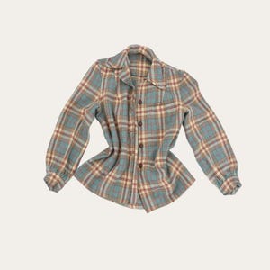 Puede incluir: Una camisa de cuadros de manga larga de estilo vintage en tonos azul, crema y burdeos. La camisa presenta un cuello clásico, una tapeta abotonada en la parte delantera y puños abotonados. La tela tiene un aspecto suave y ligeramente desgastado.