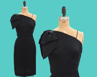 May include: A black one-shoulder dress with a large bow on the shoulder. The dress is fitted and has a knee-length hem.