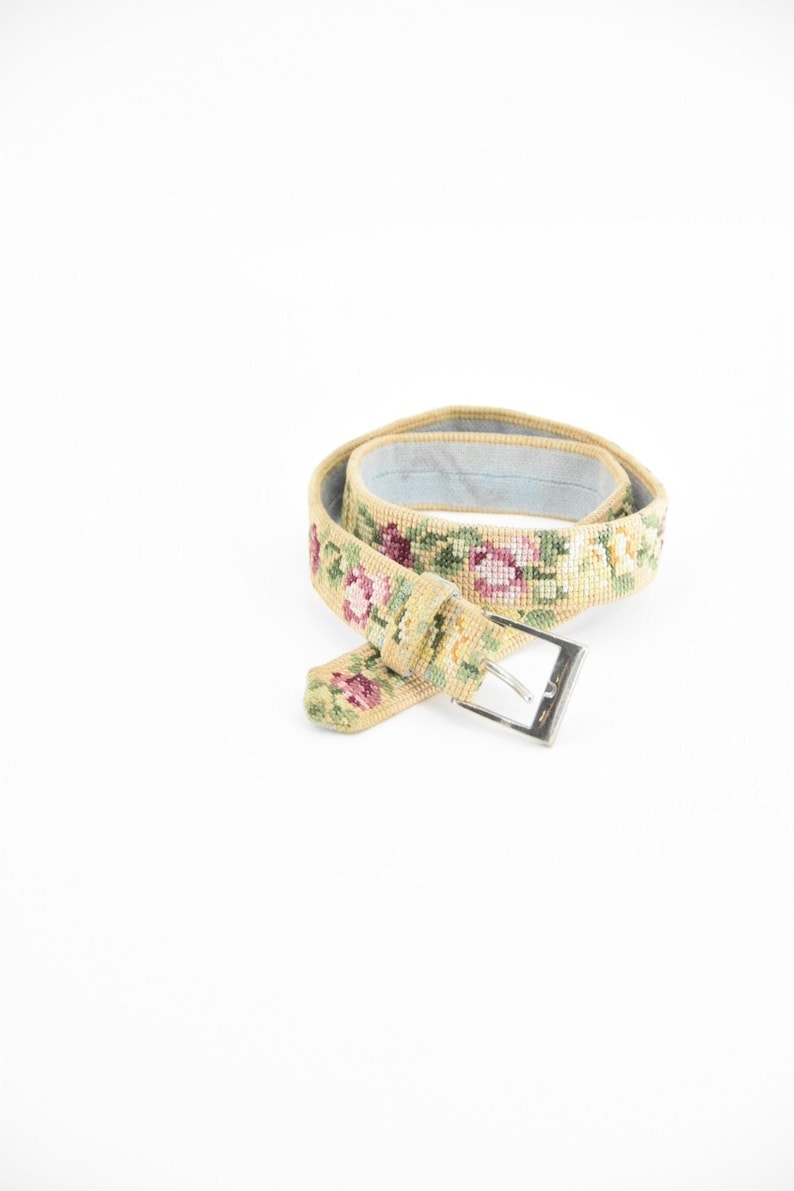 May include: A beige woven belt with a silver buckle and a floral pattern. The belt is embroidered with pink, green, and yellow flowers.