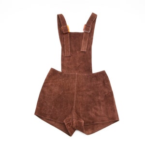 May include: Brown suede overalls with adjustable straps and buckle closures. The overalls have a short leg length and a relaxed fit.