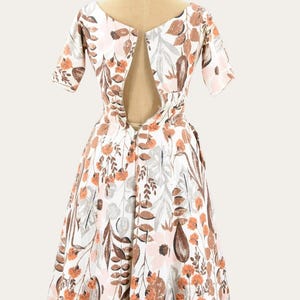 May include: A vintage white dress with a floral pattern in shades of brown, orange, pink, and gray. The dress features short sleeves and a scoop neckline with a keyhole back. The dress is displayed on a mannequin.