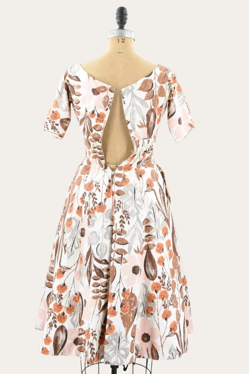 May include: A vintage white dress with a floral pattern in shades of brown, orange, pink, and gray. The dress features short sleeves and a scoop neckline with a keyhole back. The dress is displayed on a mannequin.