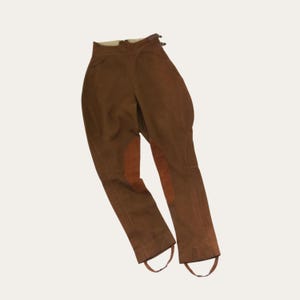 May include: Brown equestrian breeches featuring a high-waisted design with side buckle adjustments at the waistband. The pants include reinforced knee patches in a slightly darker brown tone and integrated stirrup straps at the bottom hems.