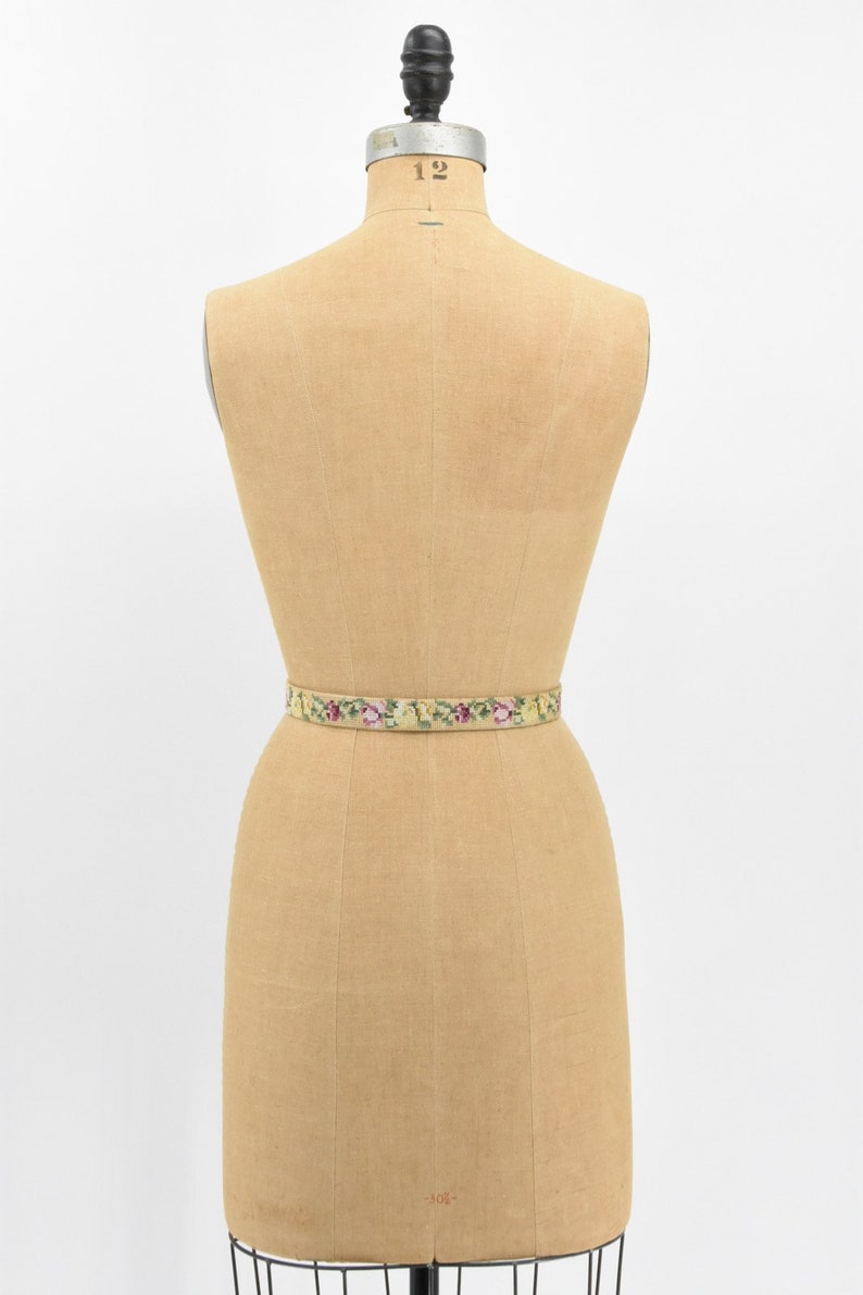 May include: A vintage floral embroidered belt in shades of green, yellow, and pink on a dress form.