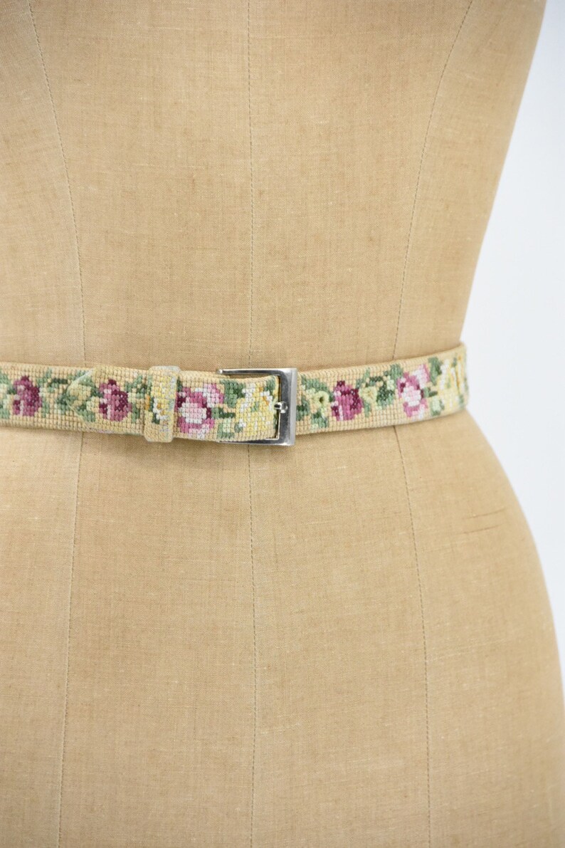May include: A beige woven belt with a silver buckle and a floral pattern of pink, yellow, and green flowers.