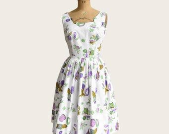 May include: A vintage white dress with a scoop neckline and a flared skirt. The dress features a repeating pattern of purple, green, and gold stylized vegetables and stars. The dress is displayed on a mannequin.