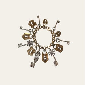 May include: A charm bracelet featuring a gold-toned chain with various charms. The charms include keys and lock pendants in gold, silver, and bronze tones. The bracelet is displayed against a white background.