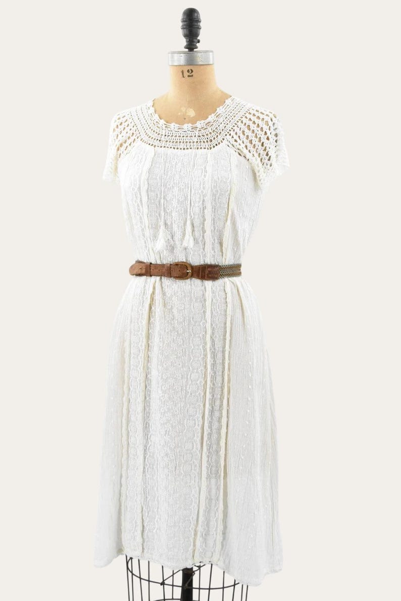 May include: A white vintage dress with a crochet neckline and short sleeves. The dress features a textured bodice and a brown belt cinching the waist. The dress is displayed on a mannequin.