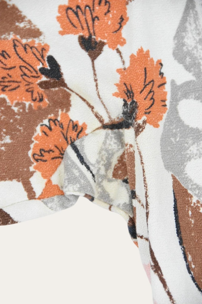 May include: Close-up of a fabric with a floral pattern. The design features orange, brown, and gray flowers on a white background. The fabric appears to be soft and possibly used for clothing or home decor.