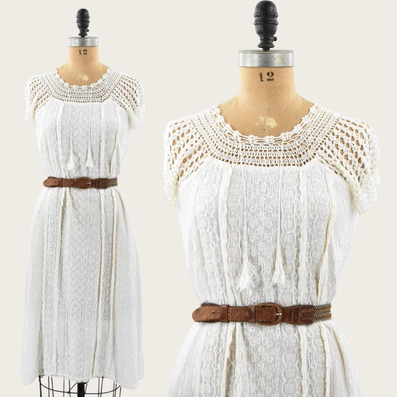 May include: A white vintage dress with a crochet neckline and short sleeves. The dress features a textured pattern and is cinched at the waist with a brown belt. The dress is displayed on a mannequin.