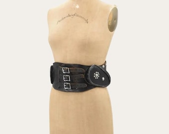 May include: A wide black leather waist belt featuring three horizontal straps with silver-toned metal buckles and a rounded side pouch adorned with a circular silver stud embellishment, displayed on a beige fabric dress form mannequin.