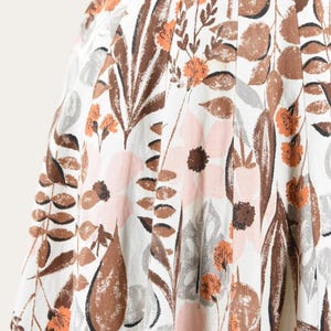 May include: Close-up of a white fabric with a floral pattern. The design features large brown leaves, pink and brown flowers, and smaller orange blossoms. The fabric has a pleated texture.