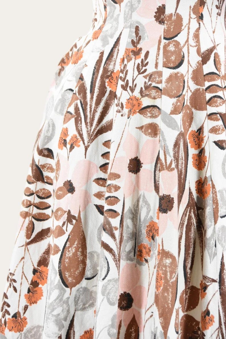 May include: Close-up of a white fabric with a floral pattern. The design features large brown leaves, pink and brown flowers, and smaller orange blossoms. The fabric has a pleated texture.